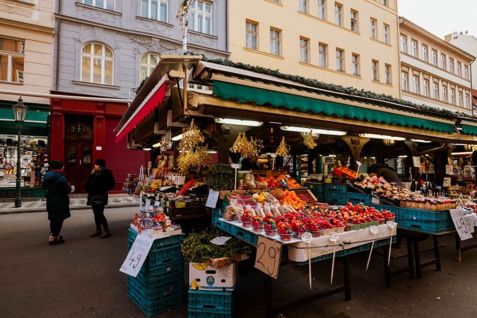An open-air market stall under a striped awning, stacked with fruit and vegetables in crates on a city street