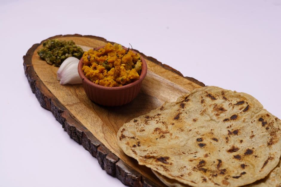 Two soft flatbreads on a wooden board next to a bowl of spiced yellow potato and a green chutney
