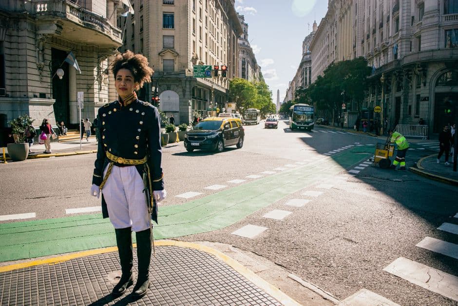 A black and yellow taxi and a city bus on a downtown Buenos Aires avenue, with the Obelisco at the far end and a performer in a historic military uniform in the foreground