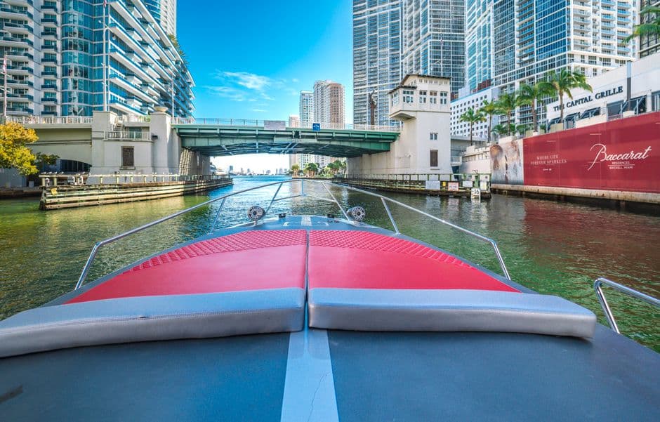 The view from the bow of a boat on the Miami River in Brickell, under a bridge between glass towers, with a restaurant sign on the right bank