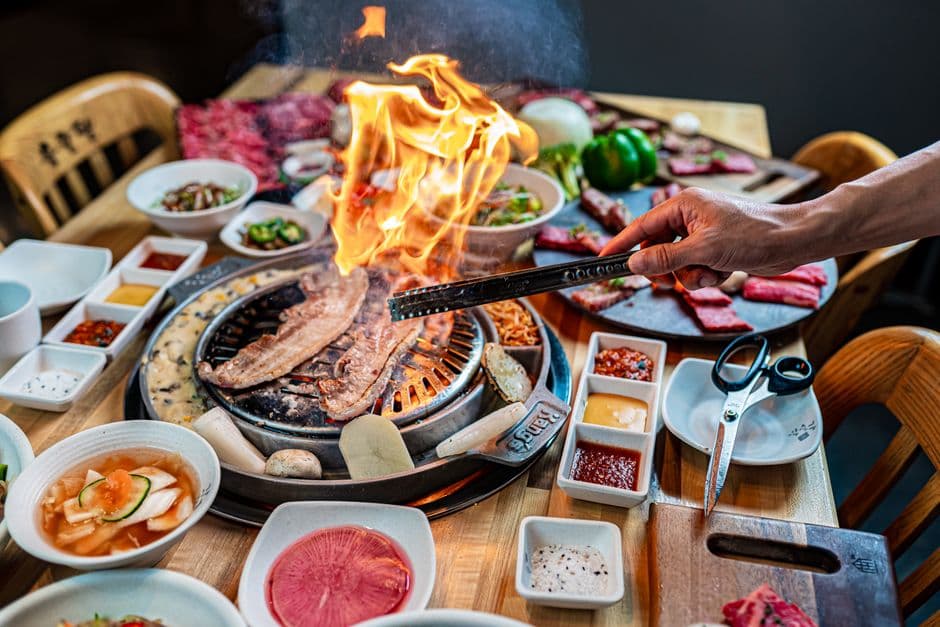 A tabletop Korean barbecue grill with pork belly cooking over an open flame, surrounded by small dishes of banchan, sauces and plates of raw marbled beef