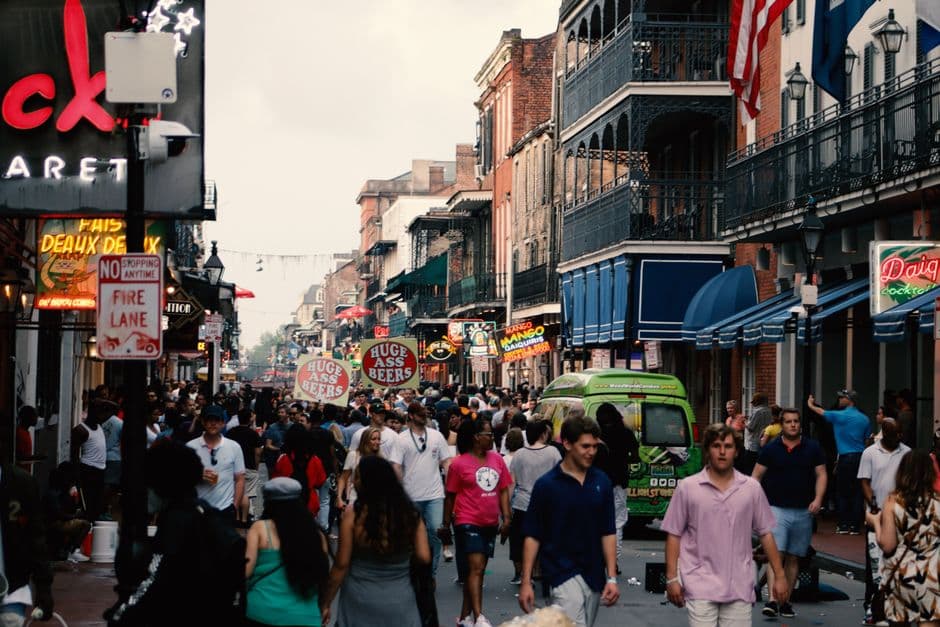 A crowded Bourbon Street in the New Orleans French Quarter at dusk, lined with daiquiri bars and neon signs advertising oversized beers, under cast iron balconies