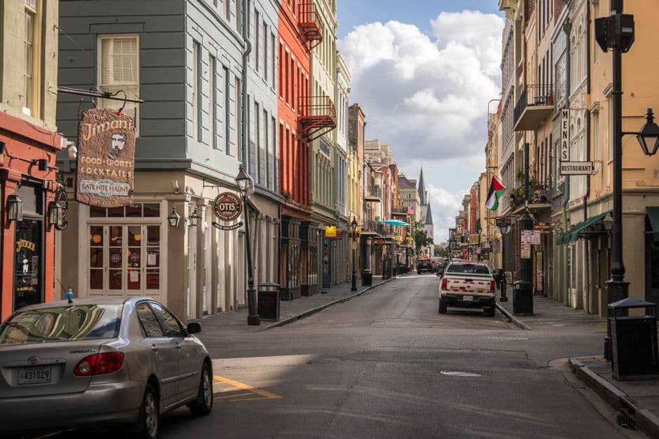 Chartres Street in the New Orleans French Quarter, with the painted storefronts and hanging signs of The Jimani and Mena's Restaurant, and the spire of St Louis Cathedral visible down the street