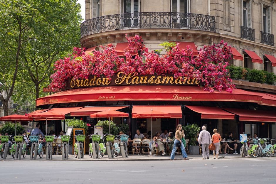 A Paris corner brasserie with red awnings, gold lettering and flower boxes, its terrace full and public bicycles parked outside