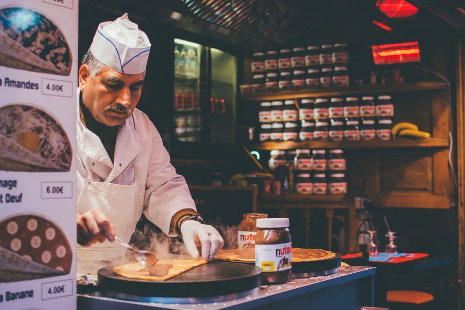 A cook in white spreading batter across a crepe griddle at a stand, with a price list in euros and shelves of spread jars behind