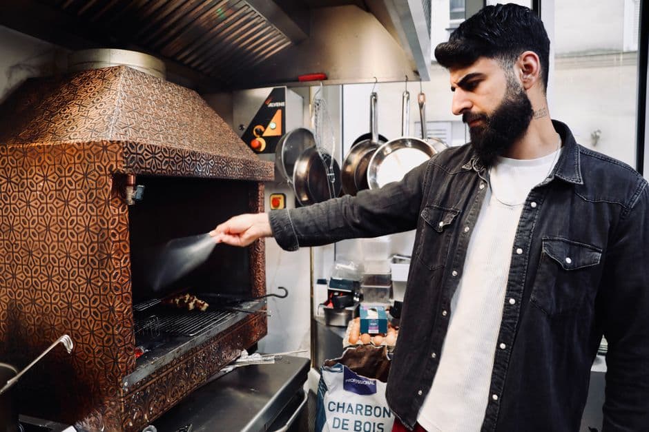 A cook reaching into a charcoal oven in a small restaurant kitchen, with a sack of charbon de bois and hanging pans beside him