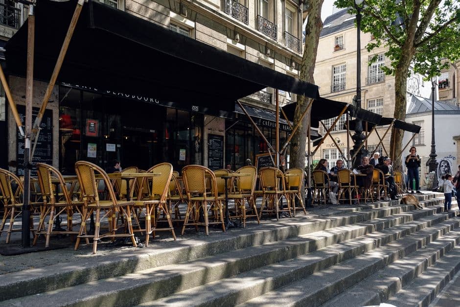 Rattan bistro chairs and tables set out along a Paris cafe terrace under a black awning, with chalkboard menus by the door