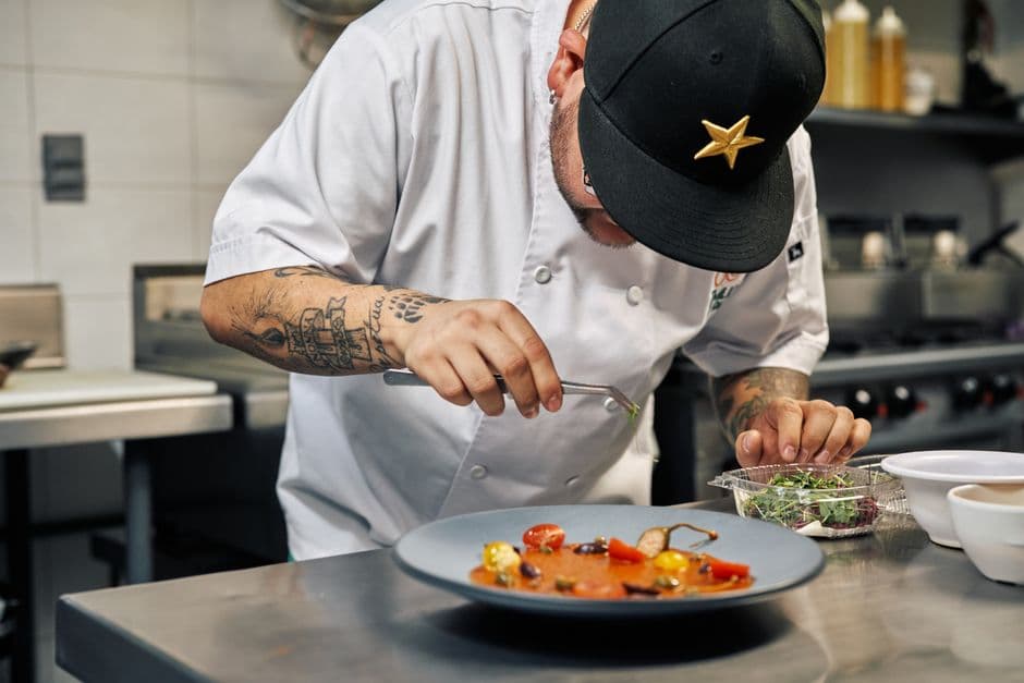 A chef in kitchen whites and a black cap using tweezers to finish a plated dish next to a punnet of microgreens