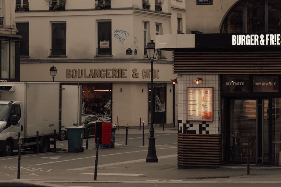A Paris street corner at dusk with a boulangerie et cantine on one side and a lit burger and fries counter opposite