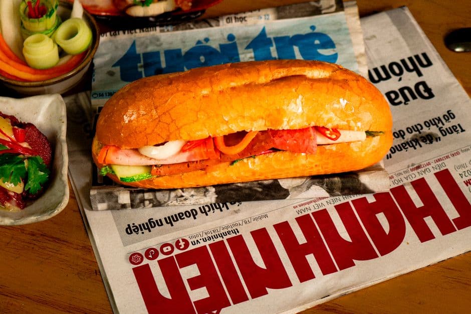 A Vietnamese banh mi on a short roll with a visibly cracked and blistered crust, filled with cold cuts, pickled carrot, cucumber, chili and cilantro, resting on Vietnamese newspapers with dishes of pickles beside it