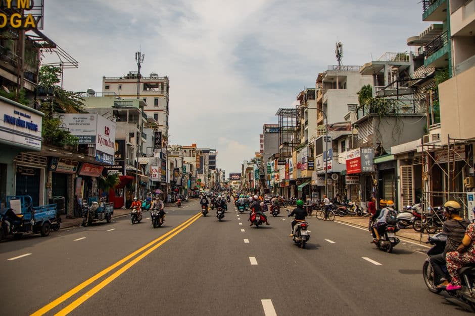 A wide Ho Chi Minh City street busy with motorbikes, lined with narrow mid-rise buildings and Vietnamese shopfront signage under a bright hazy sky
