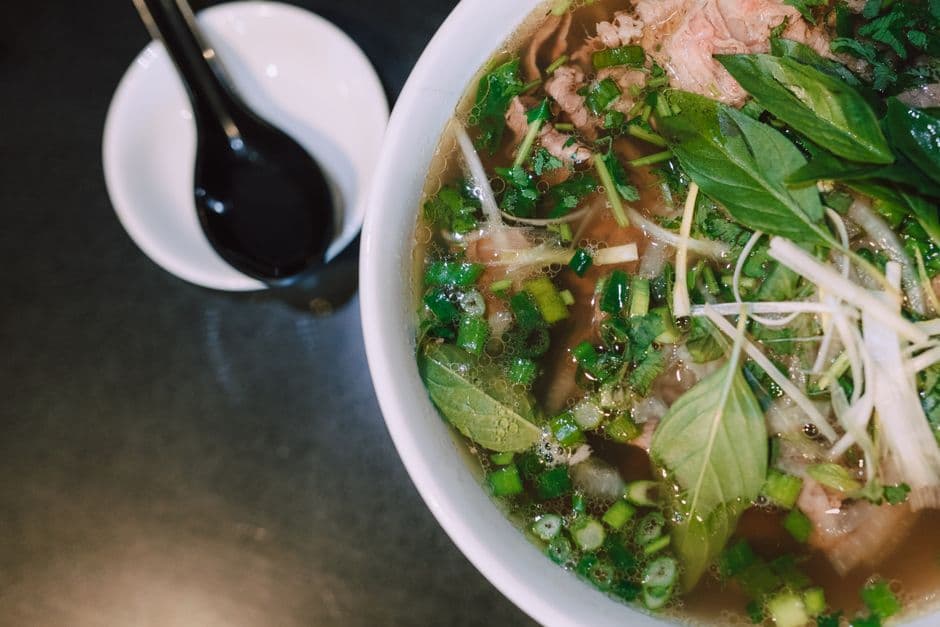Overhead view of a bowl of pho with clear broth, rice noodles, sliced rare beef, spring onion, Thai basil leaves and bean sprouts, with a small white dish of dark sauce and a black spoon beside it