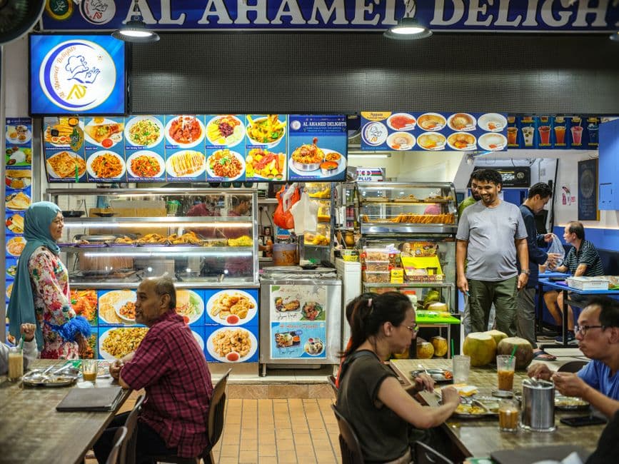 Diners at shared tables in front of an Indian Muslim food stall in a Singapore hawker center, with photo menus above a glass display counter