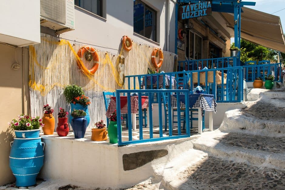 A traditional Greek taverna terrace with blue-painted chairs, checked tablecloths, whitewashed steps and painted pots of flowers