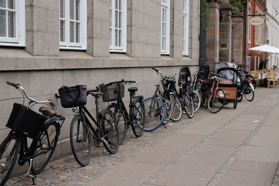 A row of bicycles parked along a cobbled Copenhagen street beside a stone building, with a cafe awning and outdoor tables further down