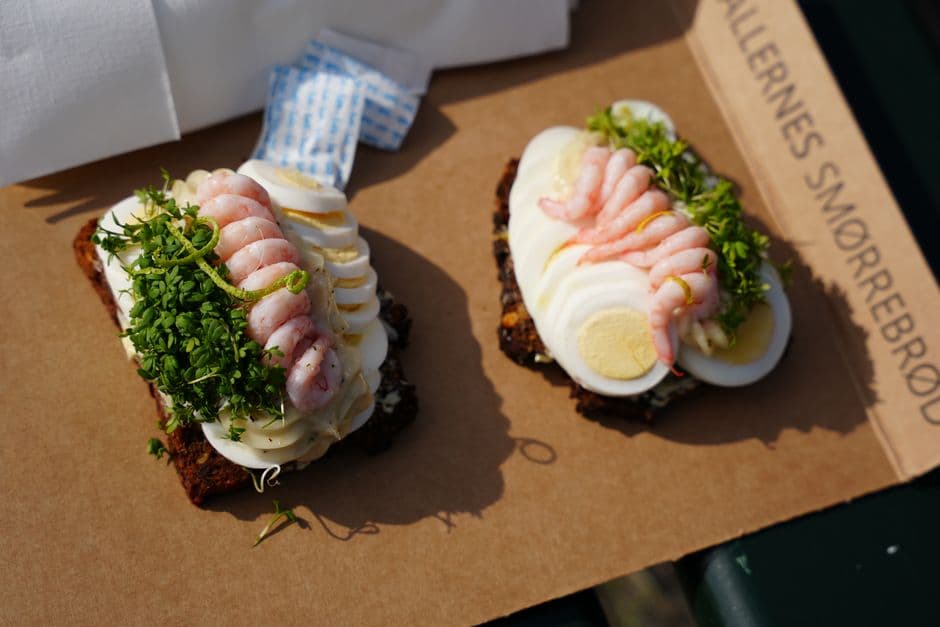 Two pieces of smorrebrod topped with cold shrimp, sliced egg and cress on dark rye bread, served on a paper board from a smorrebrod counter