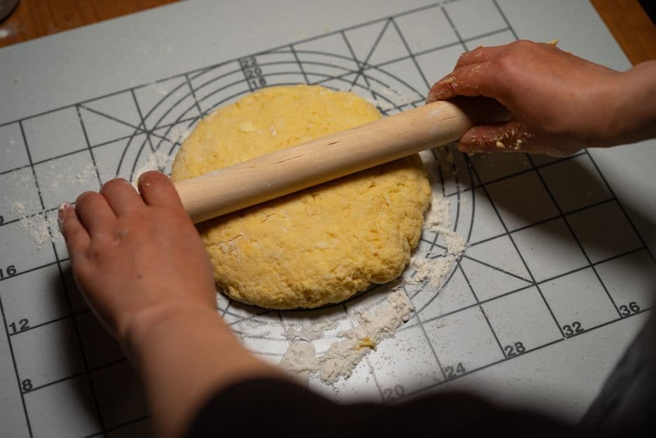 Two hands rolling out a disc of yellow egg pasta dough with a long wooden pin on a floured mat