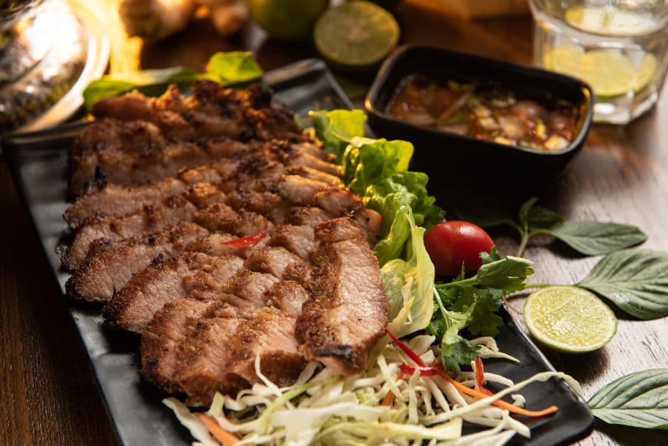 Sliced grilled pork neck on a black plate with Thai holy basil, lime, shredded cabbage and a dark chili dipping sauce