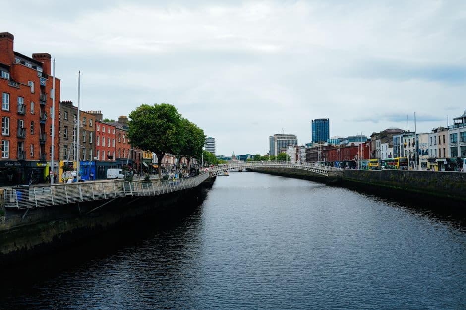 The River Liffey in Dublin under a gray sky, with red-brick buildings along the quay on the left and the white arch of the Ha'penny Bridge in the distance