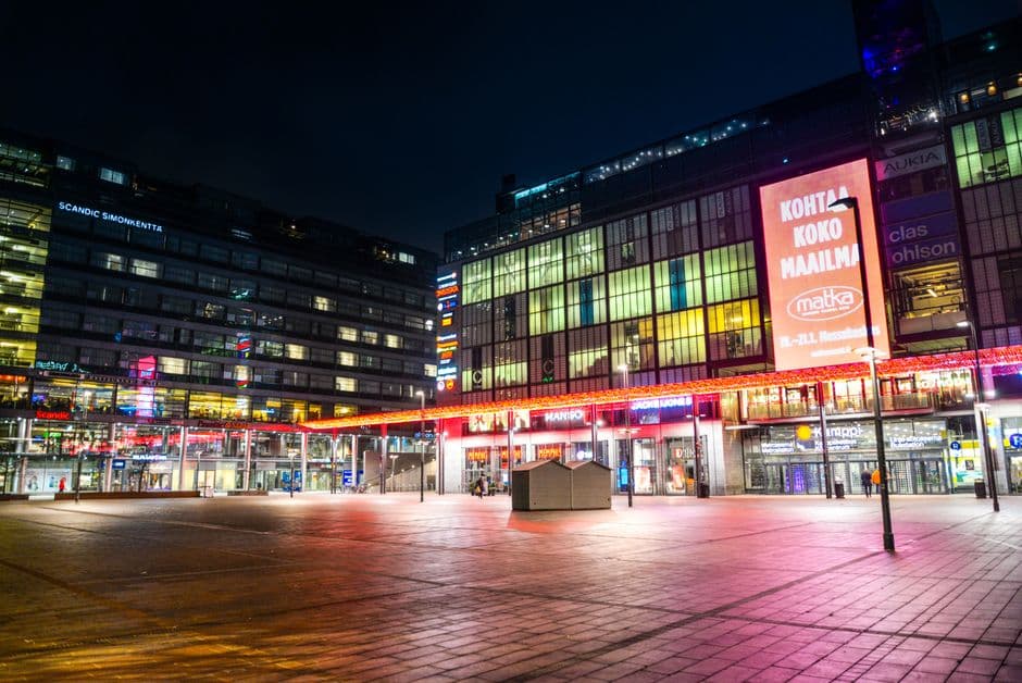 Kamppi square in Helsinki at night, lit red and pink, with the Scandic Simonkentta hotel sign on the left and a large Finnish language advertisement reading Kohtaa koko maailma on the right