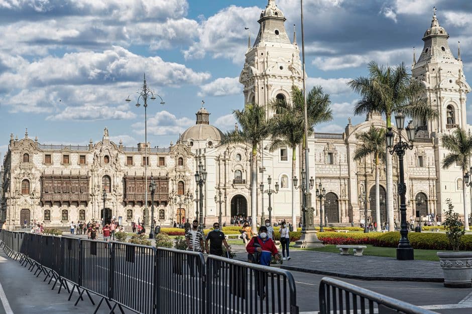 The twin-towered Cathedral of Lima and the carved wooden balconies of the Archbishop's Palace on the Plaza Mayor, with palm trees and people walking through the square