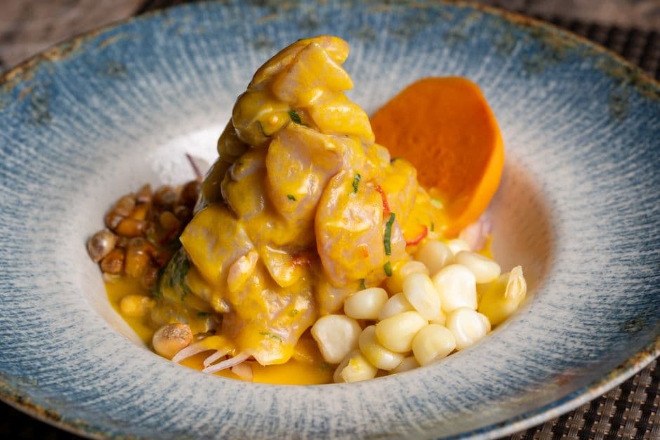 A mound of ceviche in a yellow chili sauce on a blue ceramic plate, with a slice of orange sweet potato, a pile of large white corn kernels and a spoonful of toasted corn
