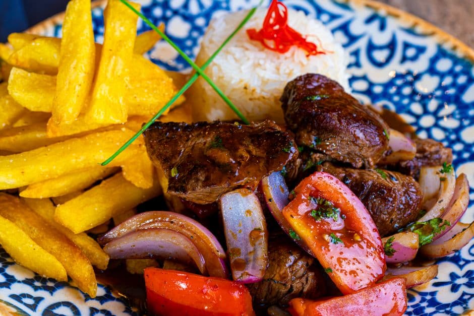Lomo saltado on a blue and white patterned plate: seared strips of beef with wedges of tomato and red onion, thick-cut fries and a mound of white rice