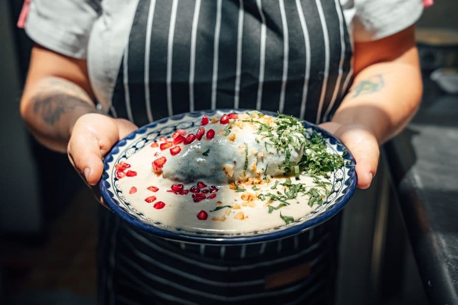 A cook holding a decorated bowl of chiles en nogada, a stuffed poblano chile under white walnut sauce scattered with pomegranate seeds and parsley