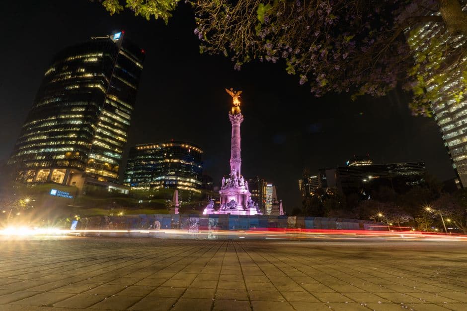 The Angel of Independence lit up at night on Paseo de la Reforma in Mexico City, with office towers behind and car light trails across the roundabout