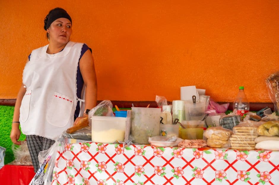 A street food vendor standing behind a table of masa, tostadas and salsas under a bright orange wall