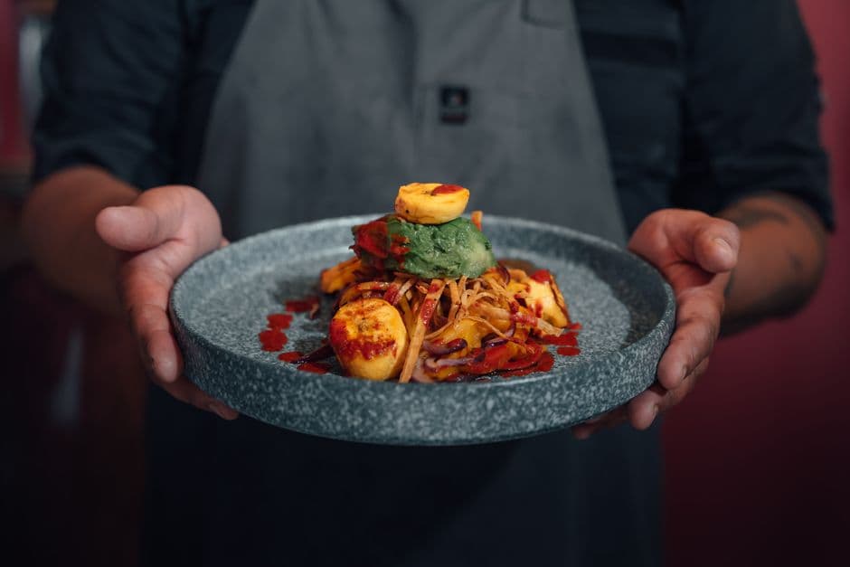 A cook holding a stone bowl with a plated modern dish of plantain, avocado, crisp strips and a red sauce