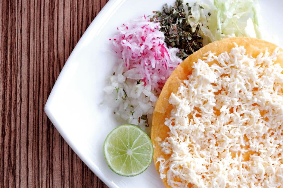 A Mexican tostada topped with crumbled queso fresco, served with lime, shaved radish, chopped onion and dried oregano on a white plate