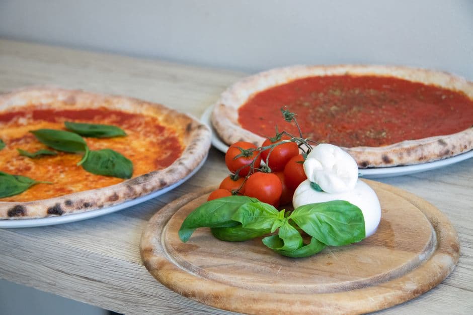 Two whole Neapolitan pizzas, a marinara and a margherita, with the raised blistered rim the specification calls a cornicione, beside fresh mozzarella, vine tomatoes and basil