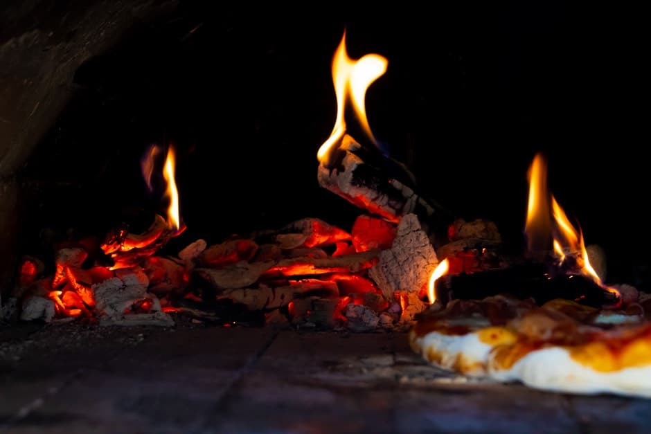 Split wood logs burning down to embers on the floor of a brick pizza oven, with a pizza cooking on the deck beside the fire