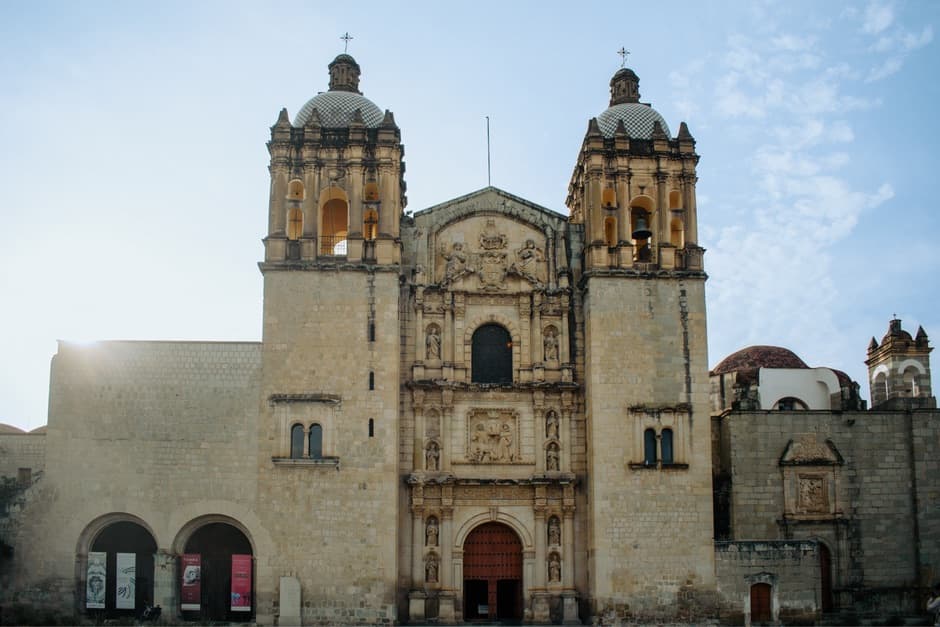 The stone facade and two bell towers of the Santo Domingo de Guzmán church in Oaxaca, each tower topped with a tiled dome, under a pale blue sky