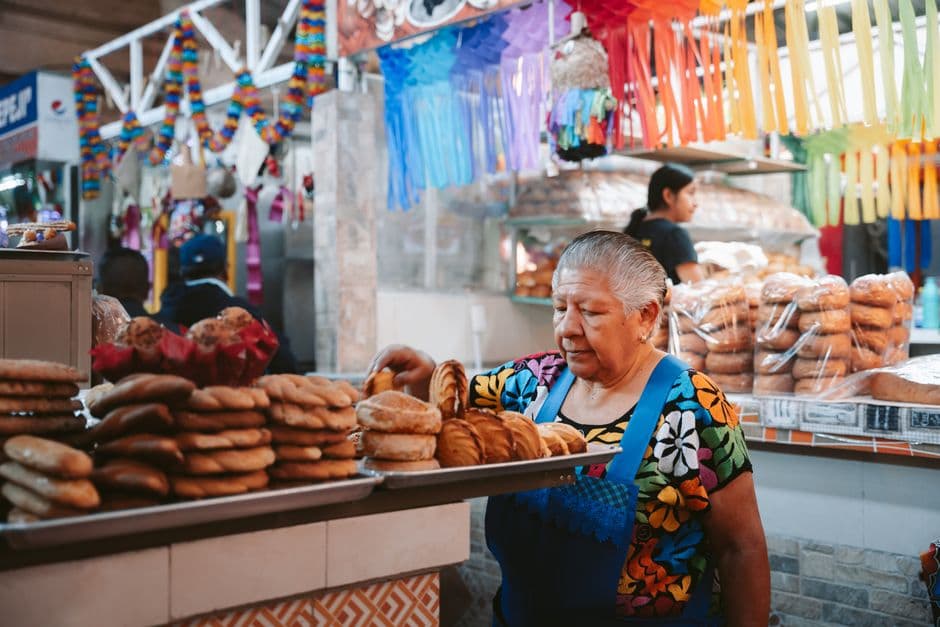 A woman in an embroidered floral blouse and blue apron arranging round loaves of sweet bread on metal trays at a market bakery stall, with bagged bread and colored paper streamers behind her