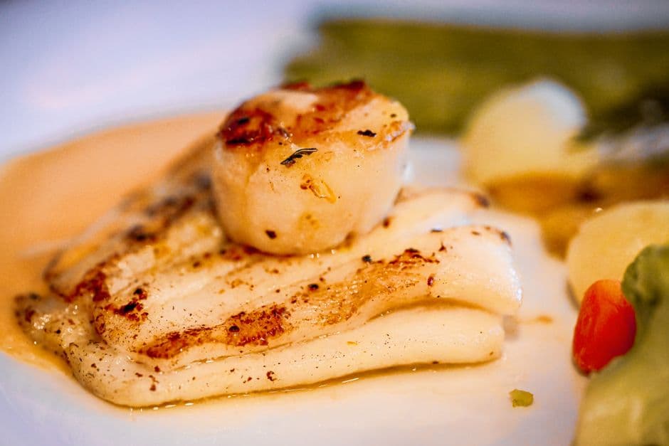 A seared scallop resting on white fish fillets with grill marks, in a pale butter sauce with green vegetables on the side