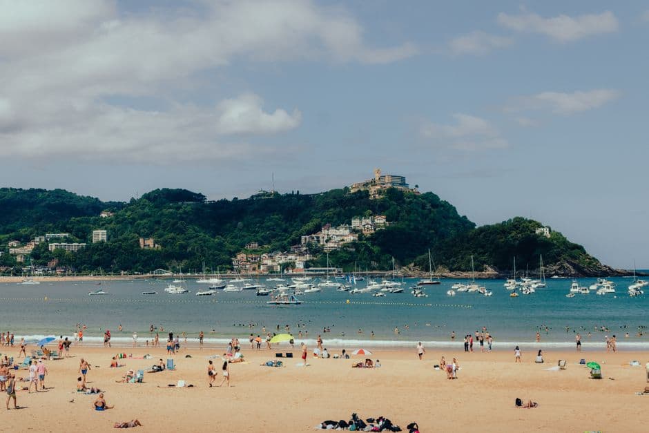 La Concha beach in San Sebastián busy with swimmers and sunbathers, with the green headland of Monte Igueldo and its hilltop building closing off the bay behind
