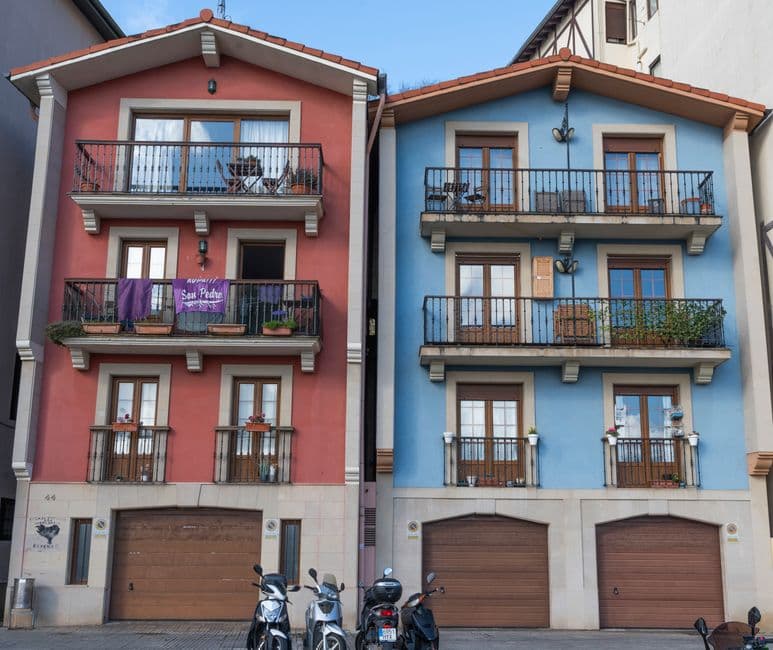 Two painted balconied townhouses in a Basque coastal town, one red and one blue, with laundry and plants on the balconies and scooters parked outside