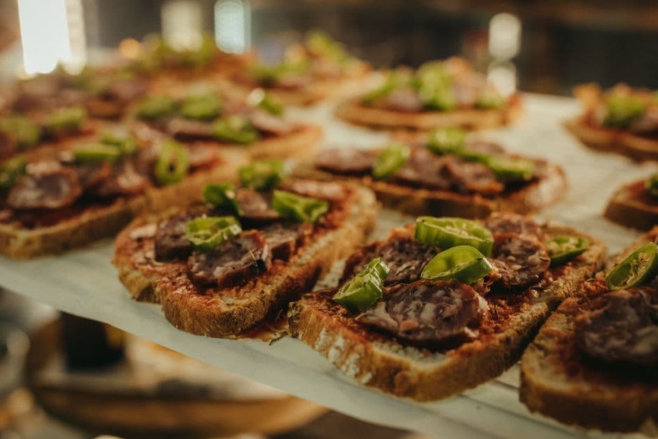 Rows of pintxos on sliced bread topped with cured sausage and green peppers, lined up on a bar counter under warm light