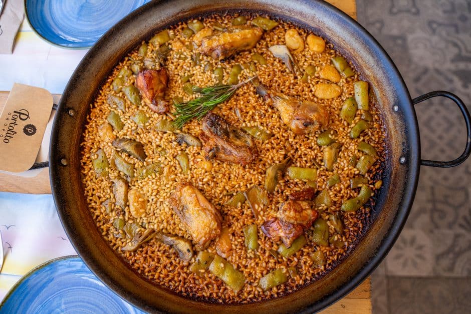 A wide shallow pan of paella valenciana seen from above, with chicken, rabbit, flat green beans, white garrofó beans and a sprig of rosemary