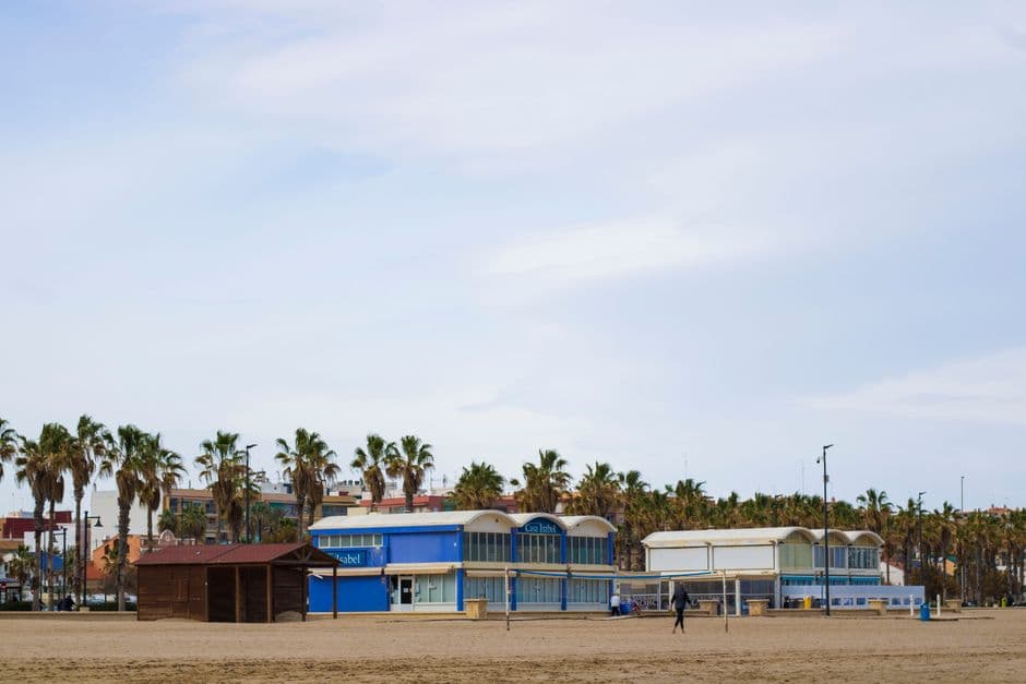 Low beachfront restaurant buildings behind the sand at Malvarrosa beach in Valencia, one painted blue and signed Casa Isabel, with a row of palm trees behind