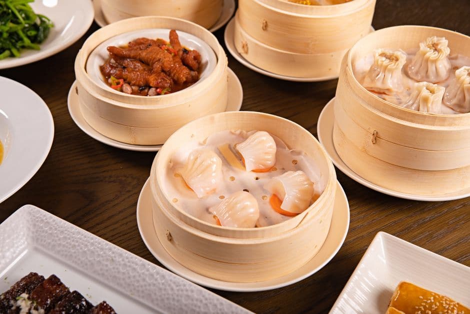 Bamboo steamers of dim sum on a wooden table, with shrimp dumplings in the center, pork dumplings to the right and braised chicken feet behind