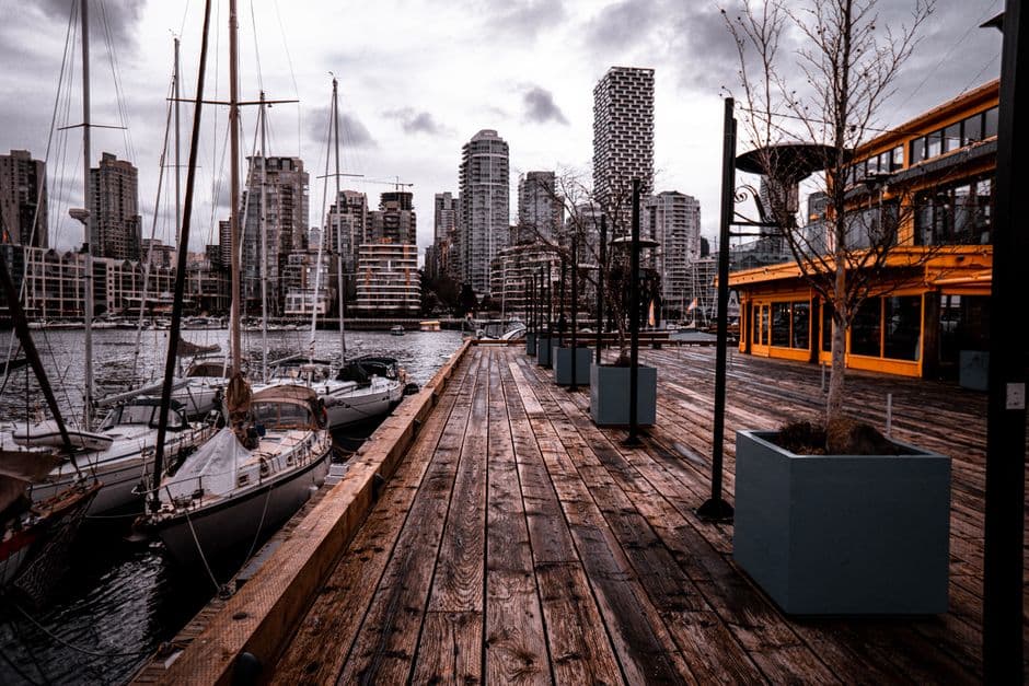 A wet wooden boardwalk on Granville Island in Vancouver with sailboats moored on False Creek and downtown apartment towers across the water
