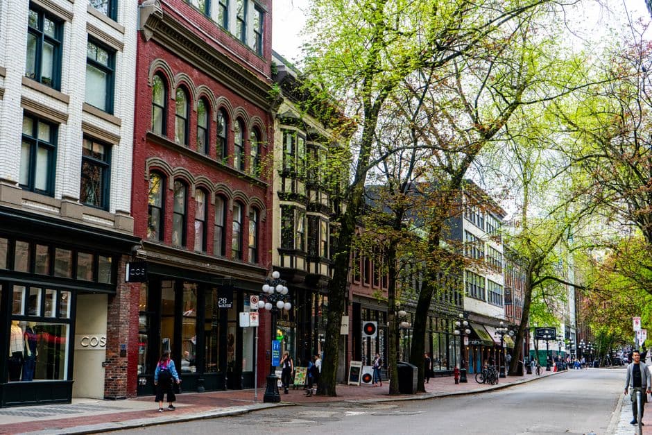 Red brick heritage buildings and spring trees along Water Street in Gastown, Vancouver, with pedestrians on the brick sidewalk
