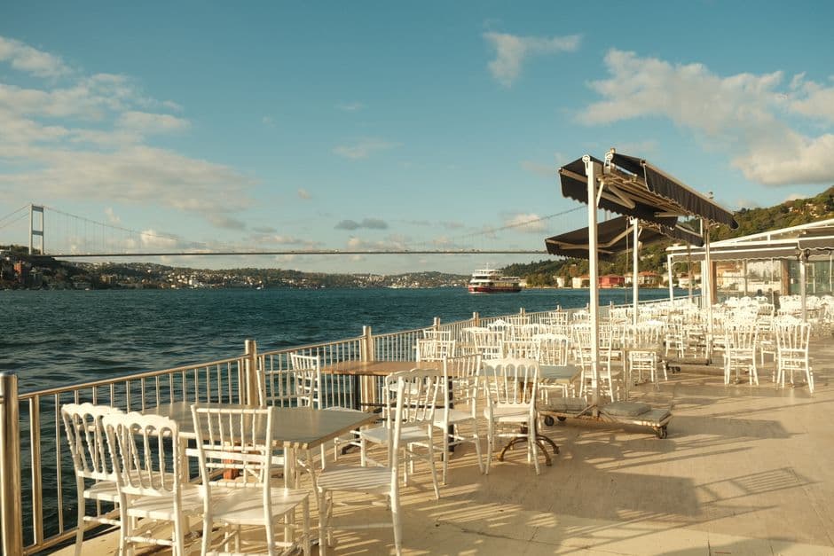 An empty waterfront restaurant terrace on the Bosphorus in Istanbul, with a suspension bridge and a passing ferry behind it