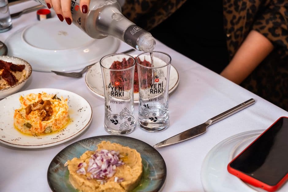 A Turkish meyhane table with raki being poured into two glasses, beside plates of hummus, haydari and other meze