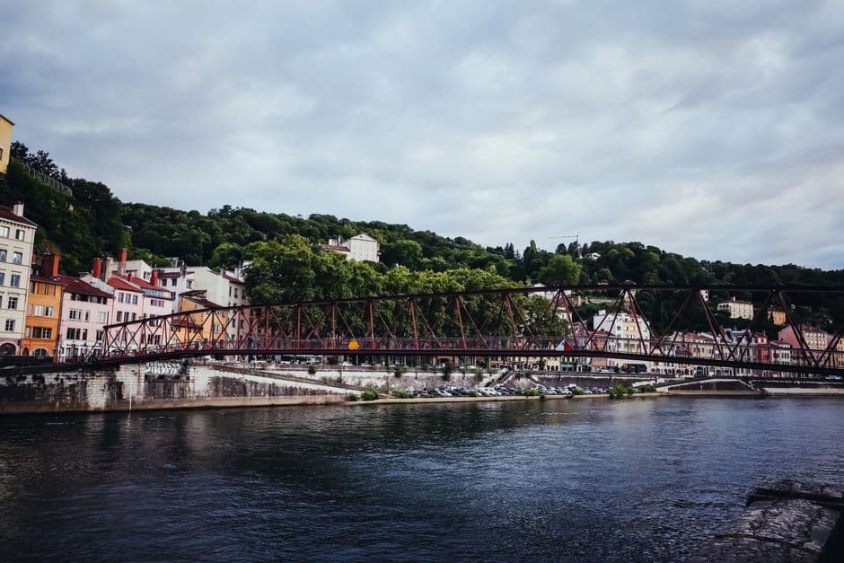 A red-painted iron footbridge crossing the Saône in Lyon, with pastel riverside buildings on the left bank and a wooded hillside behind under an overcast sky