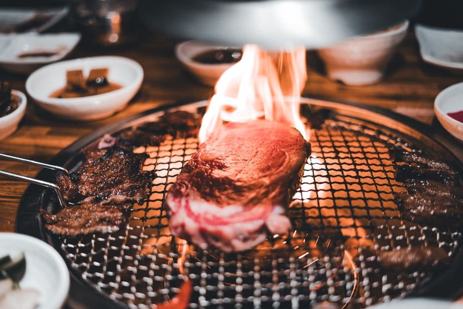 Beef and marinated short rib cooking on a charcoal mesh grill set into a wooden restaurant table, with the metal extraction hood above it and white bowls of side dishes around the rim