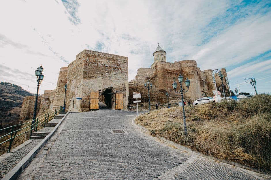 The stone walls and gate of Narikala fortress in Tbilisi with a church tower behind them, seen from the cobbled road that climbs to the entrance
